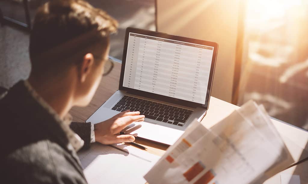 Young banking finance analyst in eyeglasses working at sunny office on laptop while sitting at wooden table.Businessman analyze stock reports on notebook screen.Blurred background,horizontal.