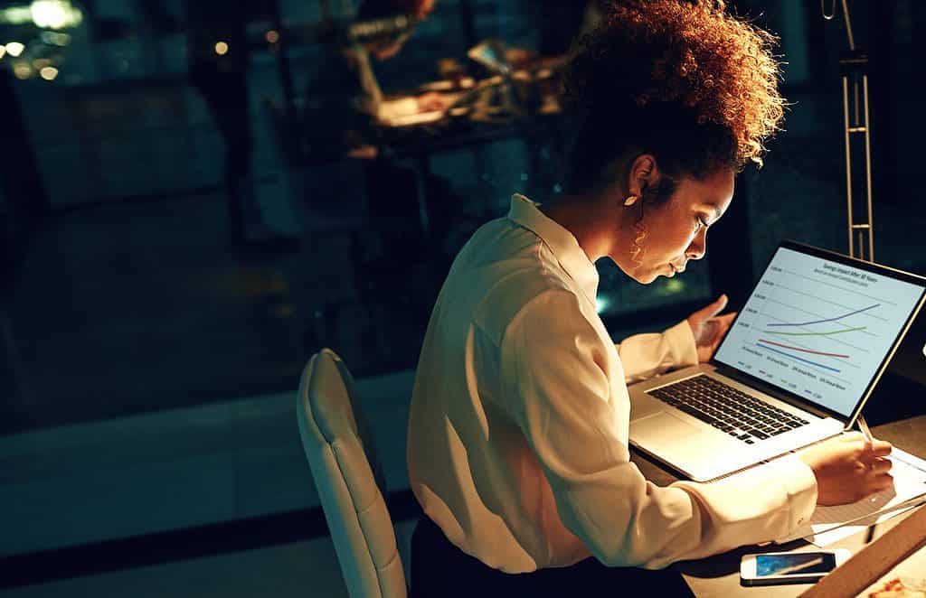 A busy woman writing down notes beside a laptop.