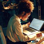 A busy woman writing down notes beside a laptop.